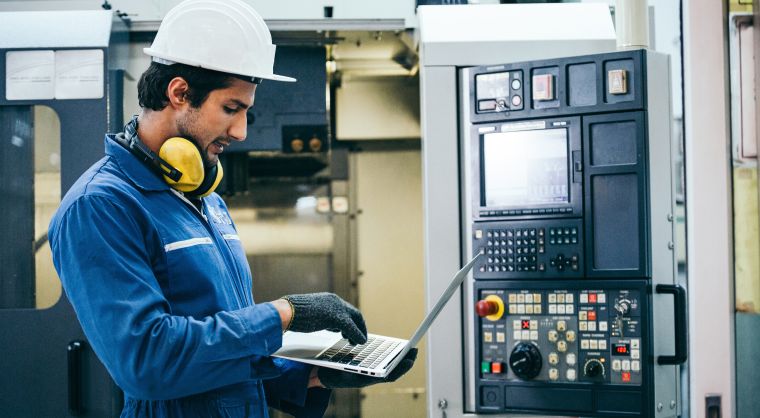 Technician in a hard hat using a laptop to perform CNC machine maintenance.