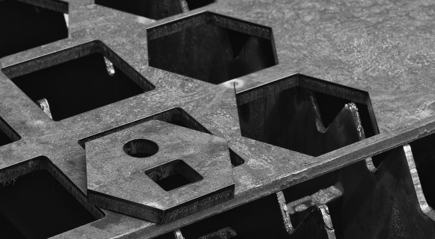Black and white close-up of CNC-cut steel plate with hexagonal cutouts and a cut part with round and square holes resting on the skeleton